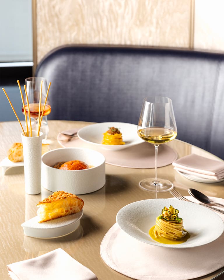 Photograph of a dish with several ingredients inside into which a hand is pouring a small bowl of sauce. The scene is completed with a glass of white wine