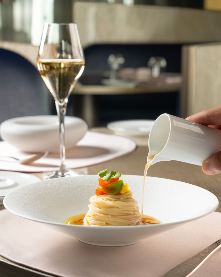 Photograph of a dish with several ingredients inside into which a hand is pouring a small bowl of sauce. The scene is completed with a glass of white wine
